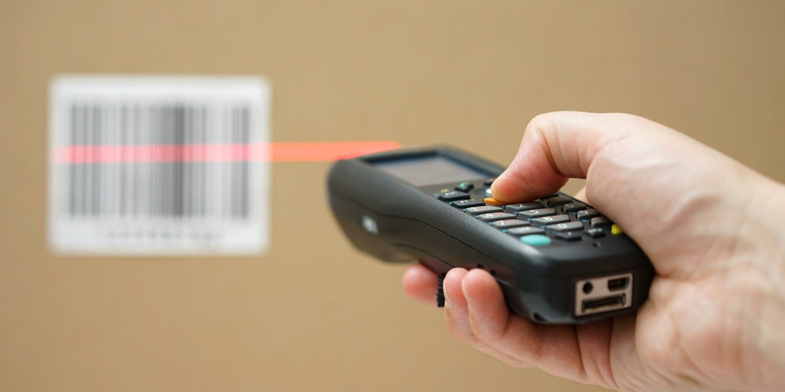 A hand holds a barcode scanner, using a red laser to scan a barcode on a cardboard box in the background.