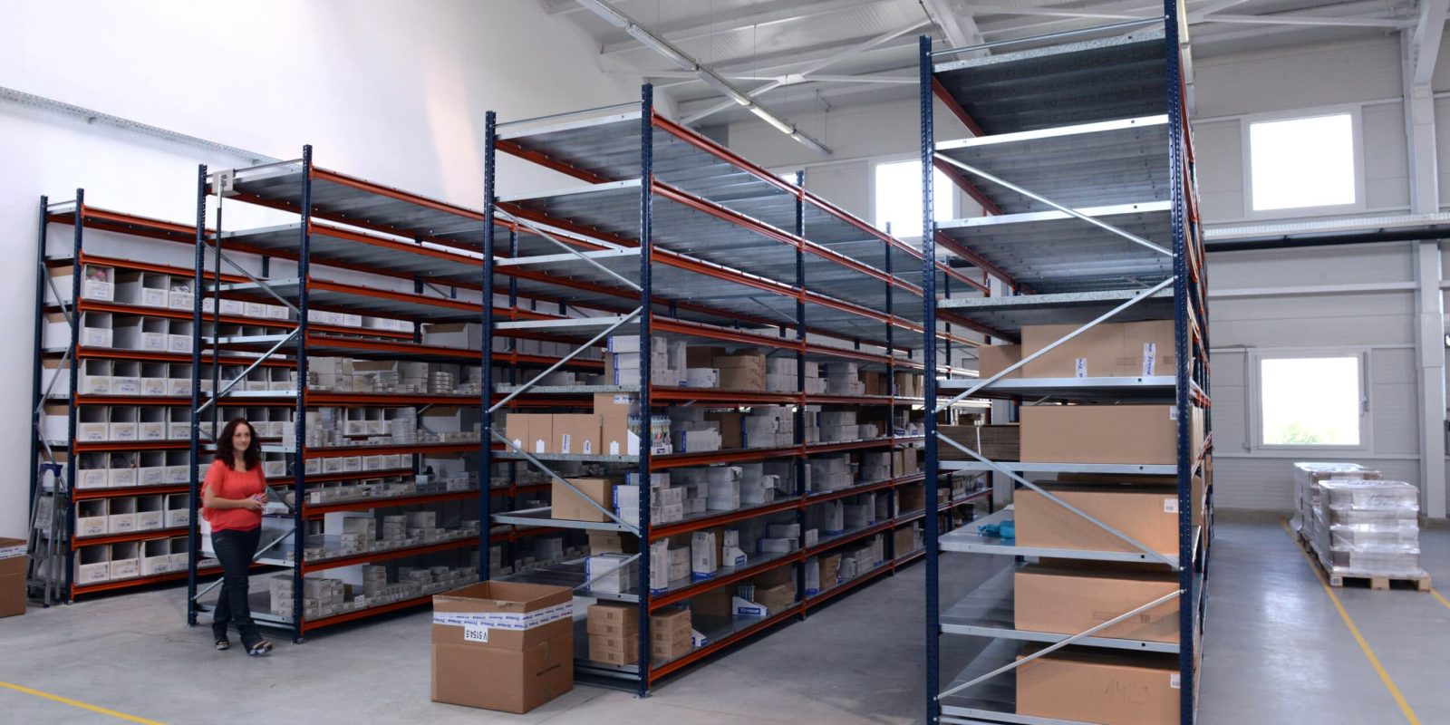 A woman walks through a spacious warehouse with tall metal shelves holding various boxes and items. The area is well-lit with high ceilings and large windows. Some boxes are stacked on the floor.