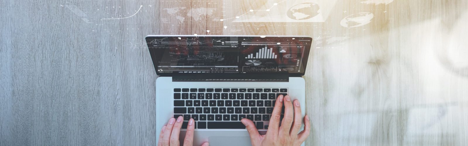 A person types on a laptop at a wooden desk, with data charts and graphs digitally overlaid above the screen, suggesting analysis or business activity. Sunlight shines on the right side of the workspace.