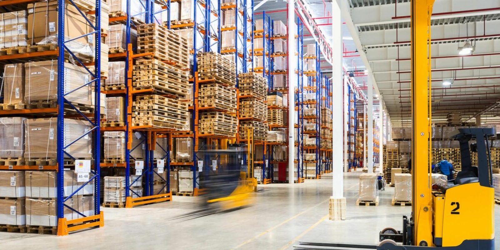 Large warehouse with tall shelves stacked with boxes, showcasing efficient slotting for organized storage. A yellow forklift is in the foreground, another in motion behind, all brightly lit with natural and artificial lighting.