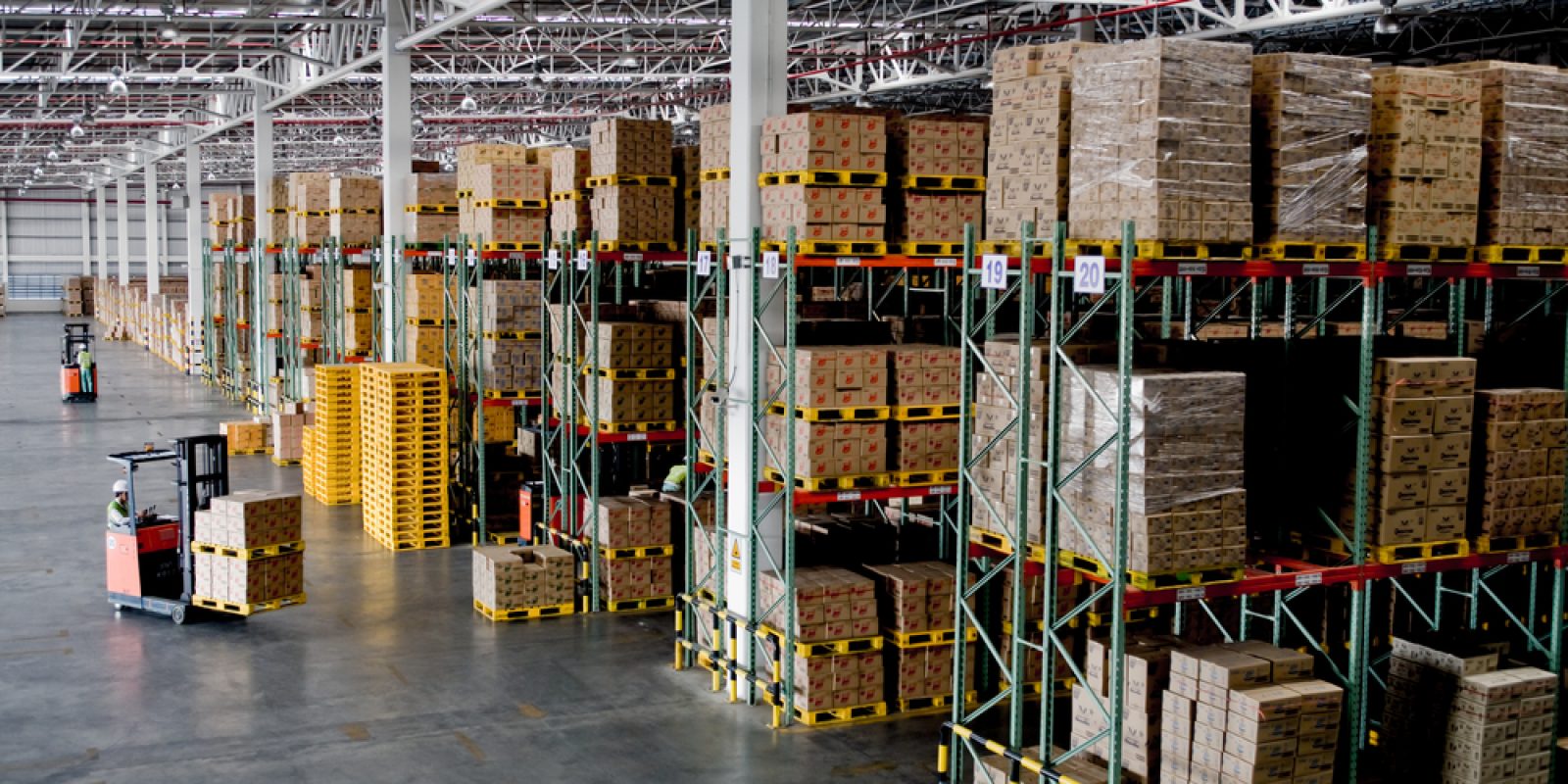 Large warehouse with high shelves stocked with boxes. Two forklifts move through the spacious, well-lit area, transporting goods. Efficient slotting ensures rows of pallets and organized inventory fill the industrial space.
