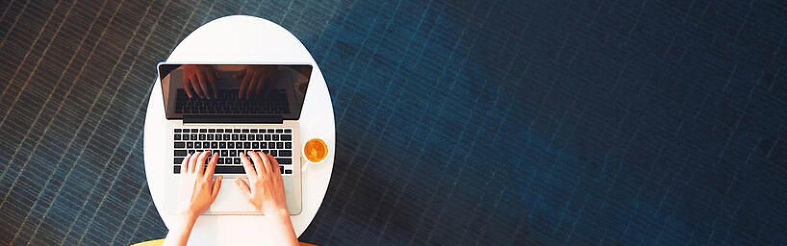 A person types on a laptop at a white round table with a cup of coffee, seated in a yellow chair on a dark blue carpet. The image is viewed from above.