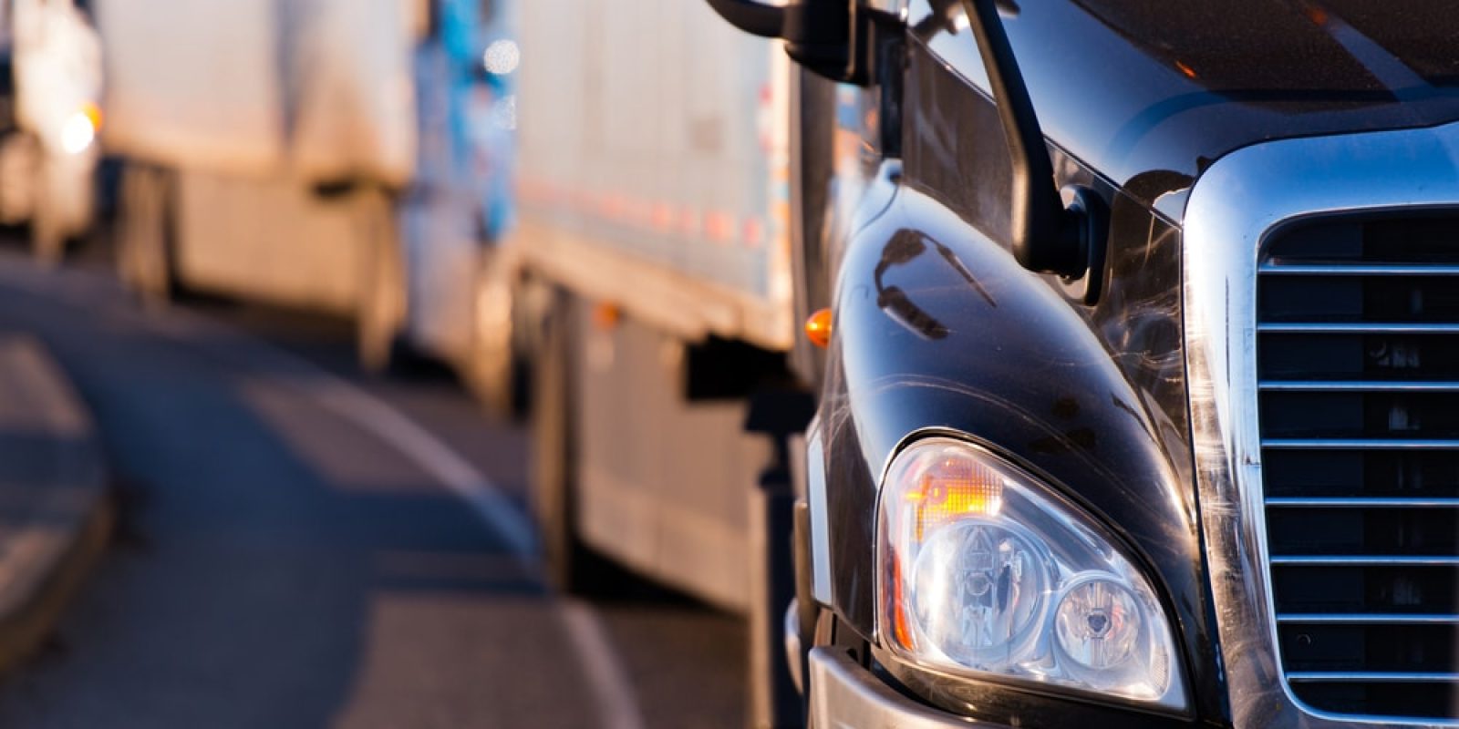 Close-up of the front of a black semi truck on a road, with other trucks lined up behind it, suggesting a convoy or traffic on a highway in the early morning or late afternoon—a vital scene in transportation vulnerable to cyber-attacks.