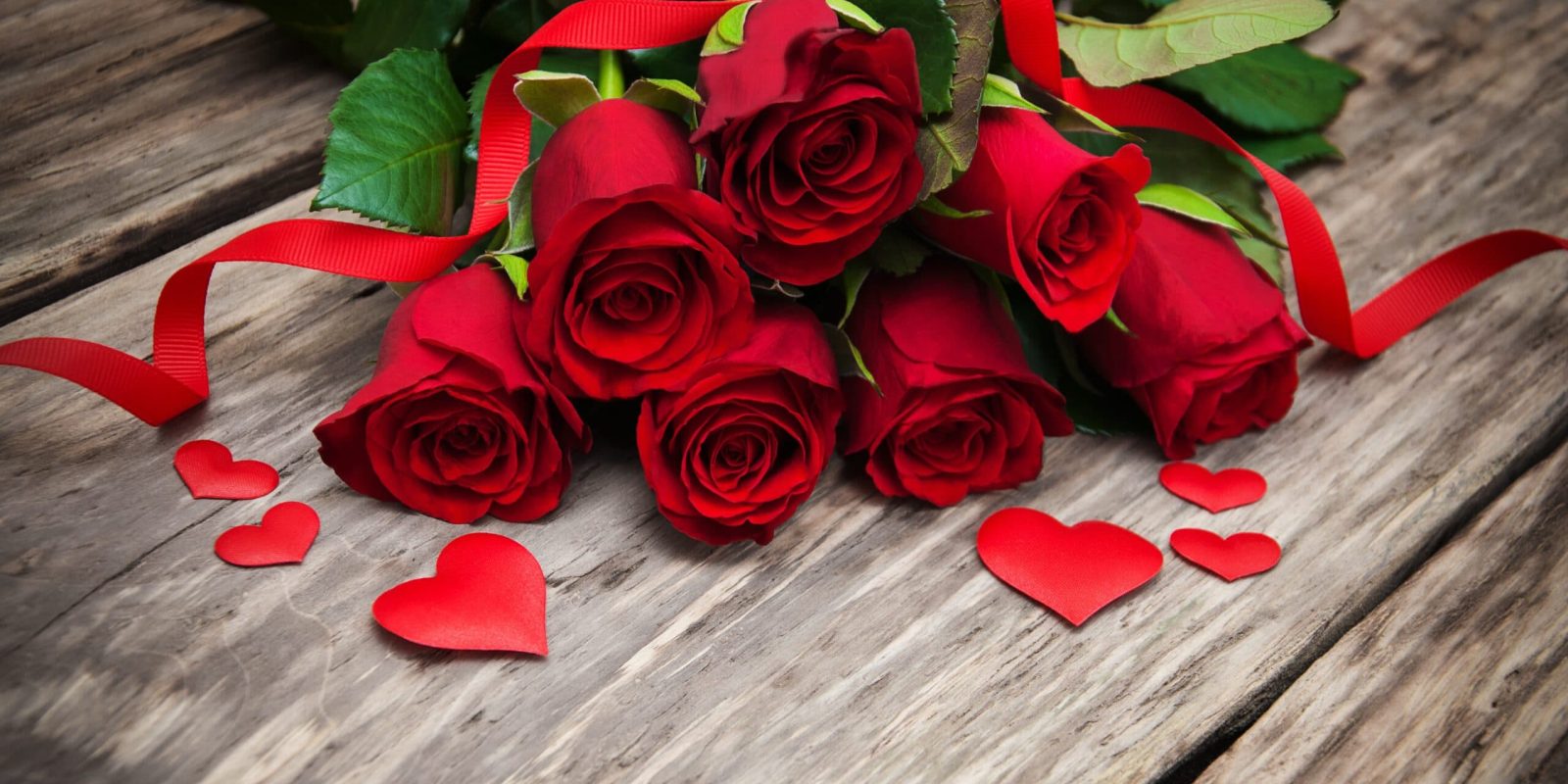 A bouquet of red roses with green leaves is placed on a wooden surface. Red paper hearts and a red ribbon are scattered around the flowers, creating a romantic atmosphere.