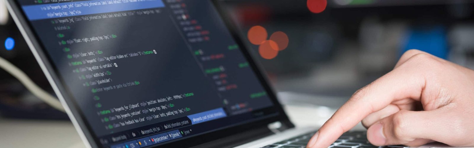 A close-up of a person’s hand typing on a laptop keyboard with programming code displayed on the screen; blurred monitors with code are visible in the background.