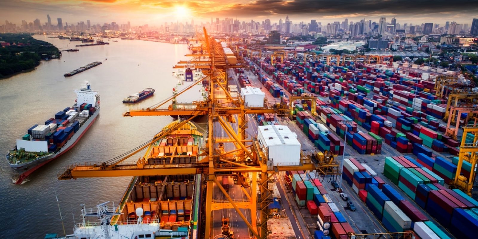 A busy shipping port at sunset with large cranes loading containers onto cargo ships. Rows of colorful shipping containers fill the docks, and a city skyline is visible in the background.