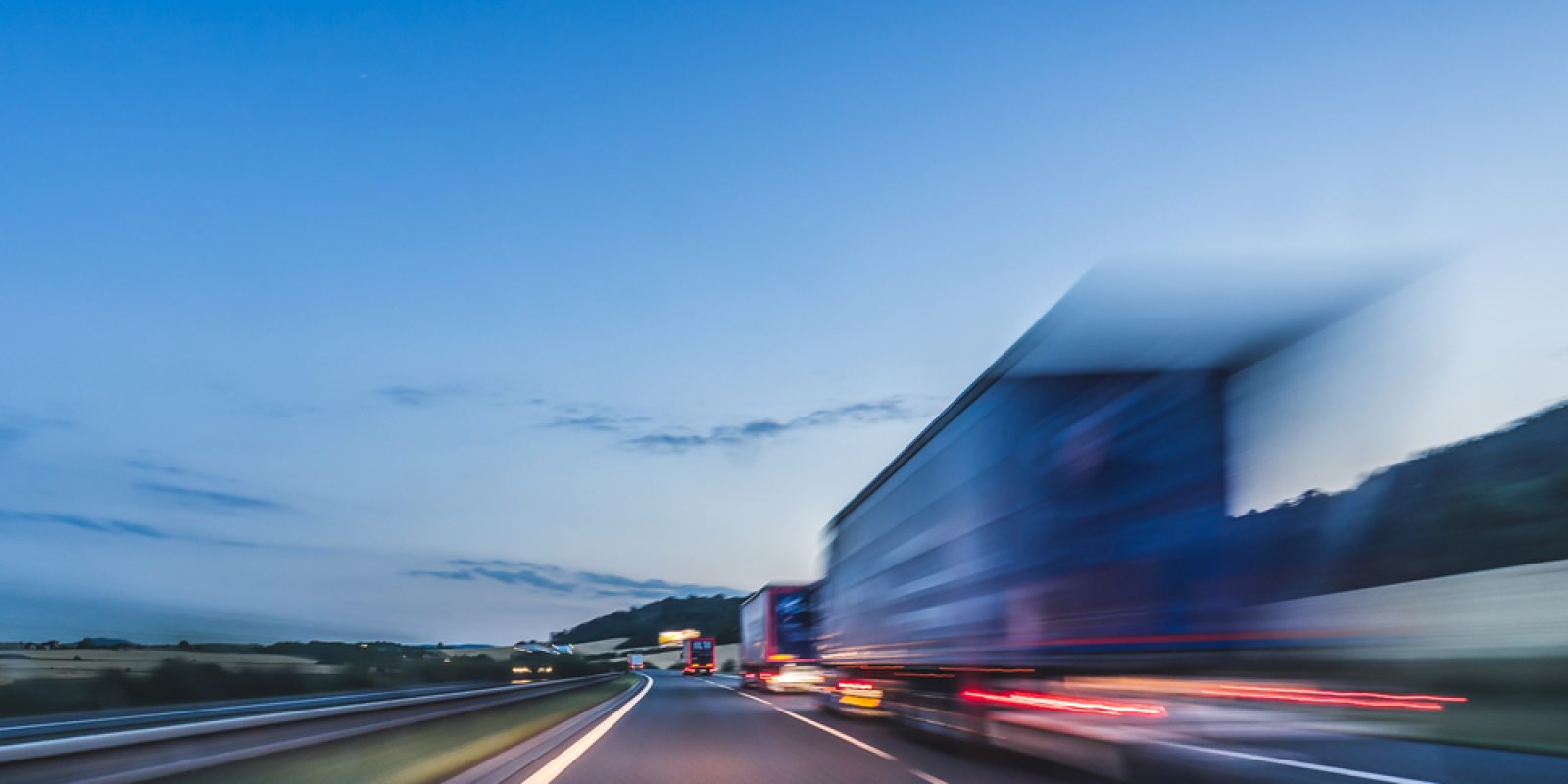 A blurred truck speeds along a highway at dusk, with a clear blue sky and distant hills visible in the background, capturing the essence of efficient transportation sourcing.
