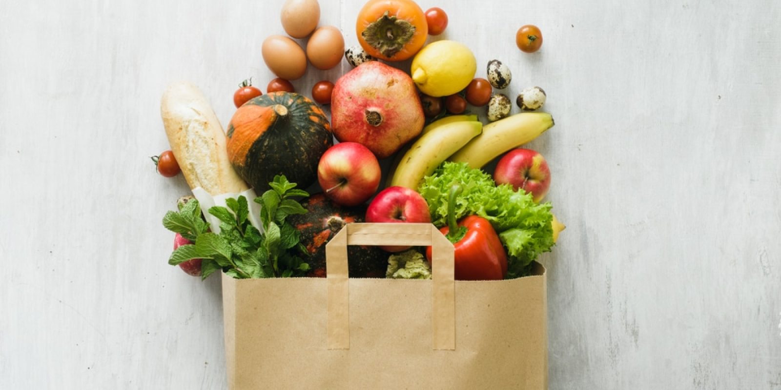A paper bag filled with various FSMA-compliant groceries, including eggs, a baguette, apples, bananas, lettuce, tomatoes, mint, a pomegranate, squash, lemons, strawberries, and cherry tomatoes on a light background.