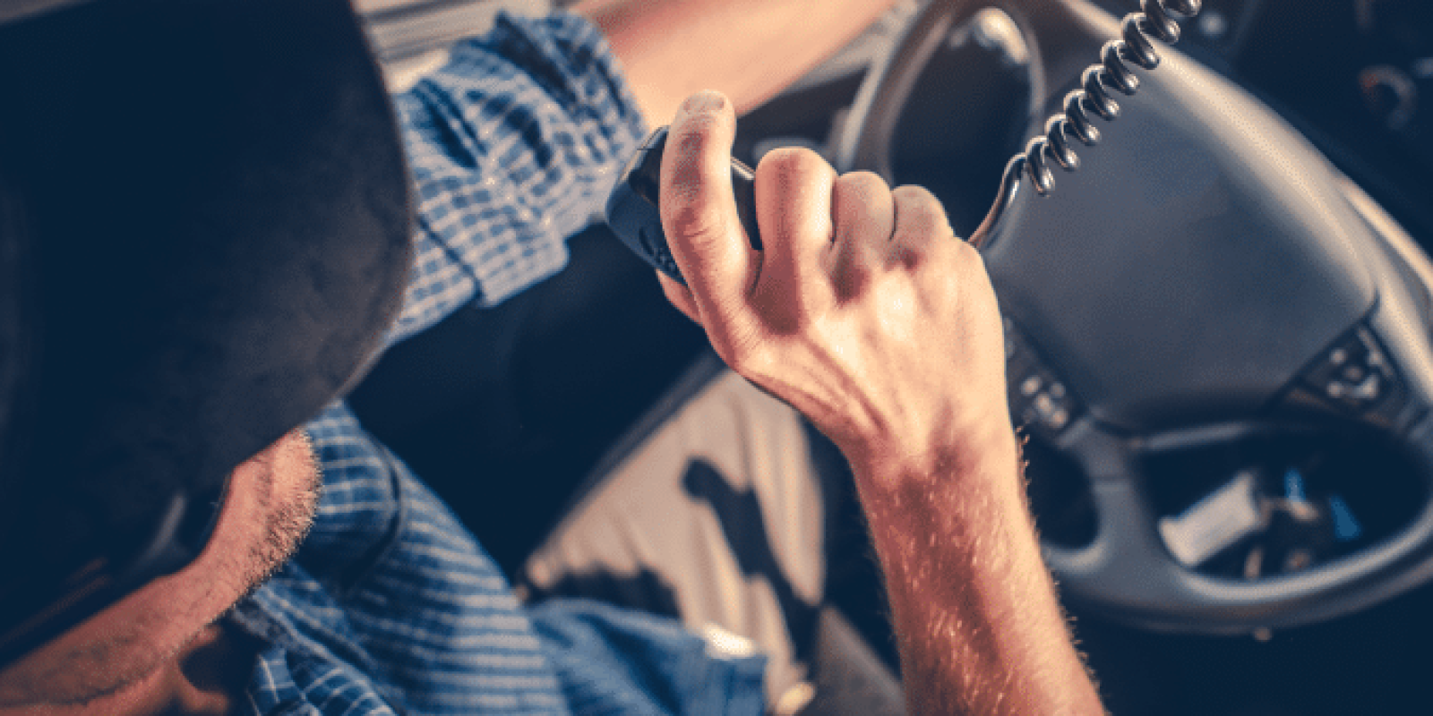 A person wearing a cap and blue plaid shirt drives a vehicle while speaking into a CB radio, with one hand on the steering wheel and sunlight streaming through the window.