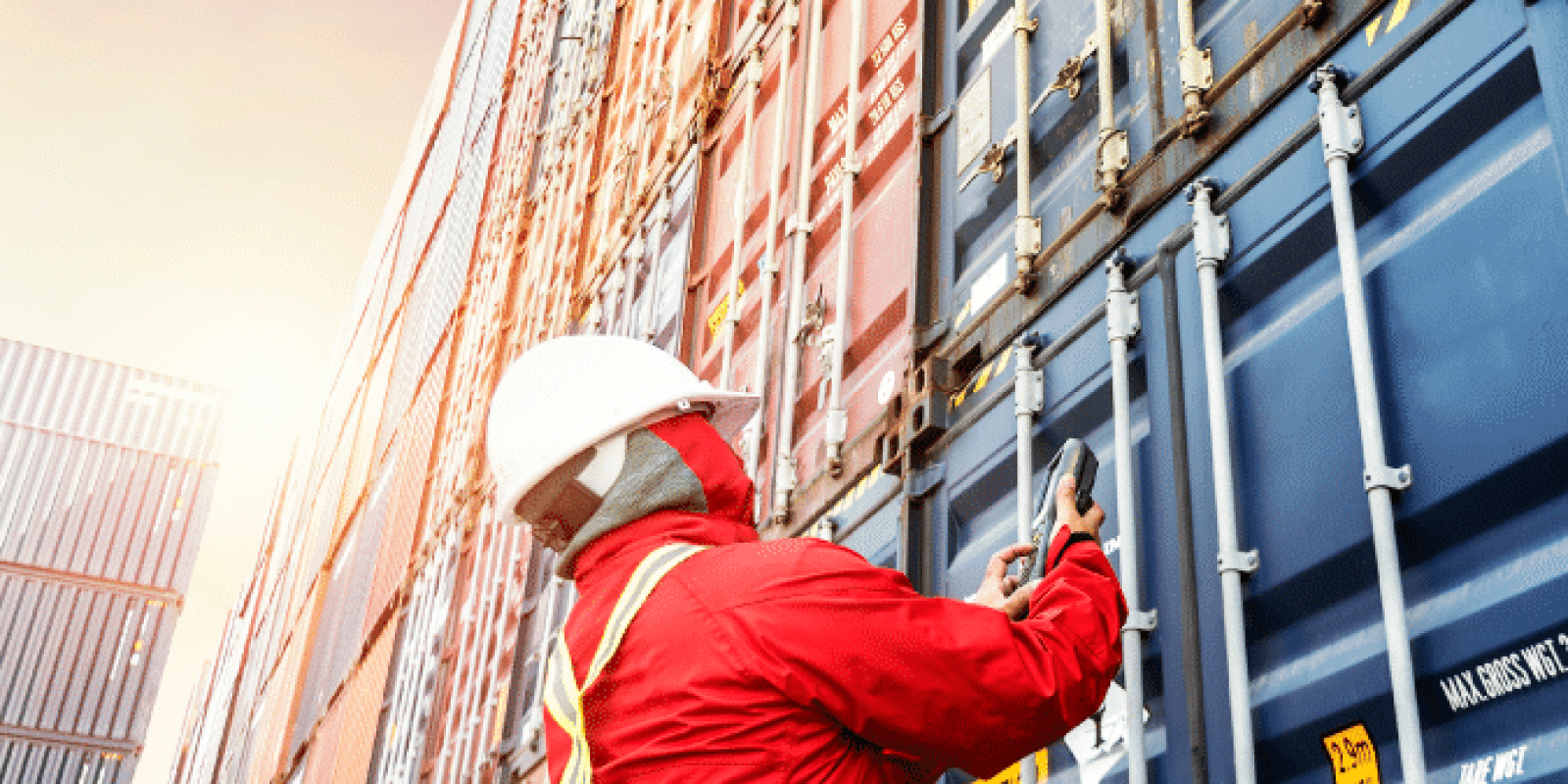 A worker wearing a red safety suit, helmet, and reflective vest inspects or secures stacks of shipping containers outdoors at a shipping yard, with sunlight illuminating the scene.