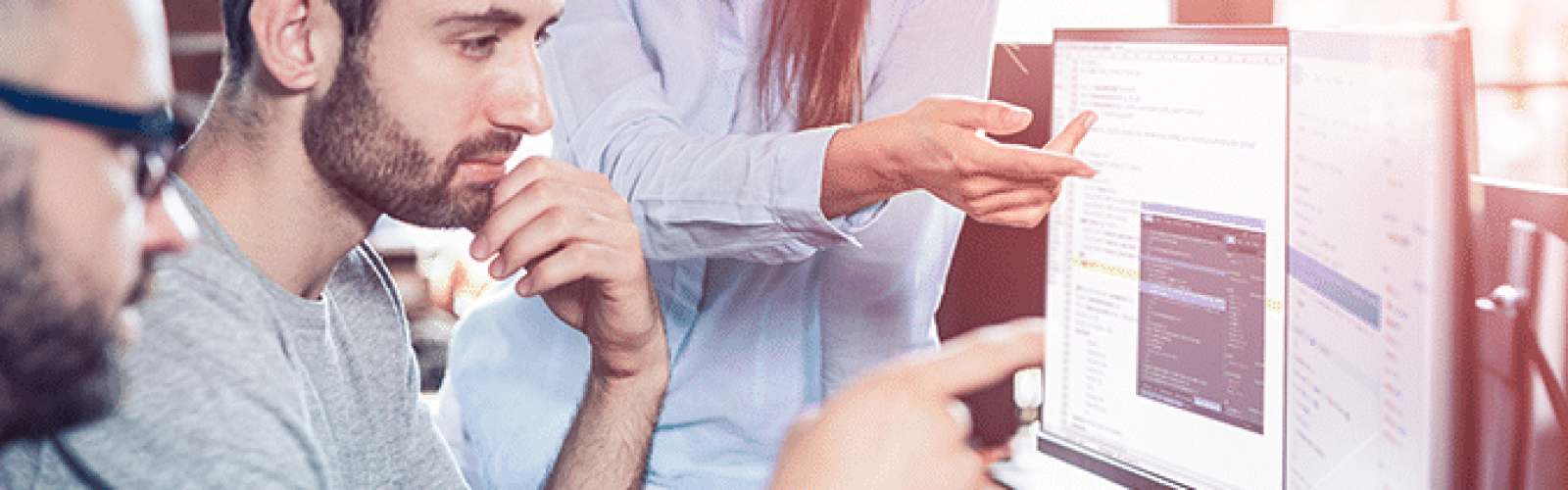 Three people collaborate in a bright, modern office. One woman stands, pointing at a monitor displaying Microsoft Power Apps, while two men sit and attentively discuss the app’s features and code on the screen.