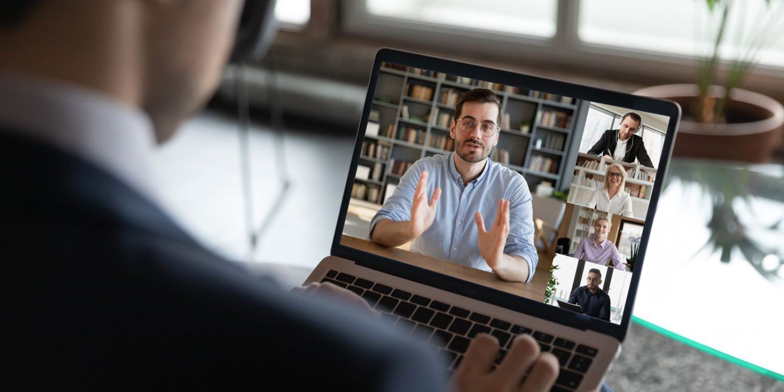 A person in a suit participates in a video conference on a laptop, with five people visible on the screen, each in their own window, engaged in a discussion. A potted plant and window are in the background.