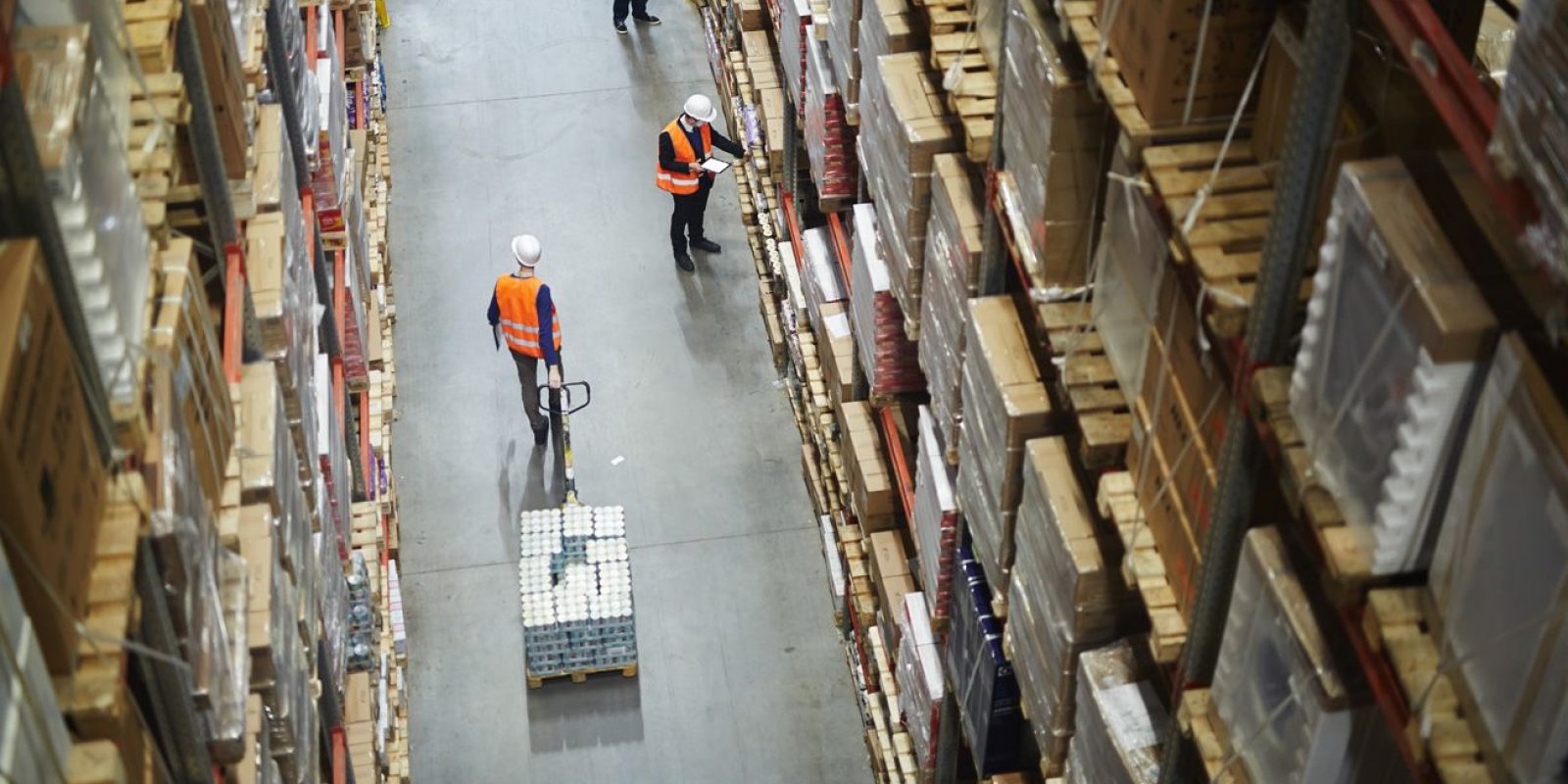 High-angle view of two workers in orange safety vests and white helmets moving and inspecting goods in a large warehouse filled with tall shelves stacked with boxes and pallets.