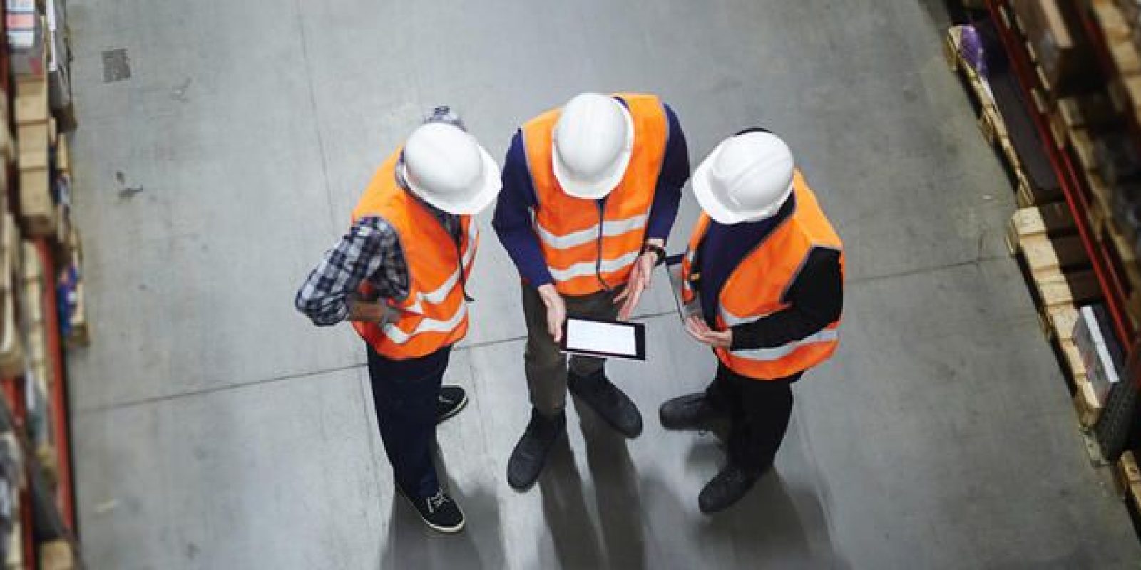 Three people wearing orange safety vests and white hard hats stand in a warehouse, looking at a tablet. The image is taken from above, showing shelves on either side and a concrete floor.