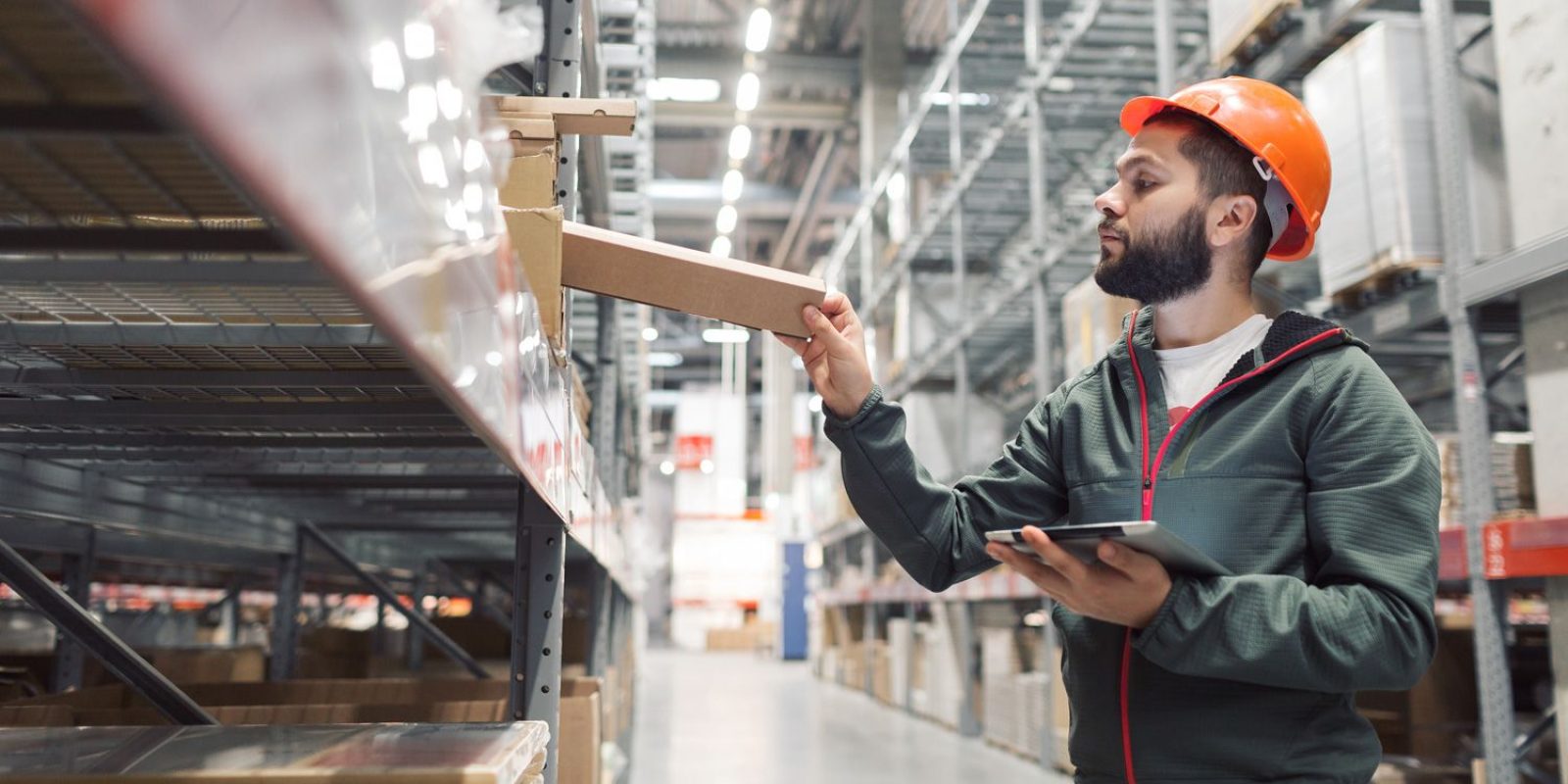 A worker wearing an orange hard hat and green jacket checks inventory on a warehouse shelf while holding a tablet, standing in a large, brightly lit industrial storage area.