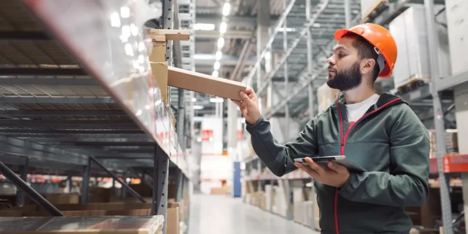A man wearing an orange hard hat and green jacket holds a tablet while placing a box on a high shelf in a large warehouse filled with shelves and storage boxes.