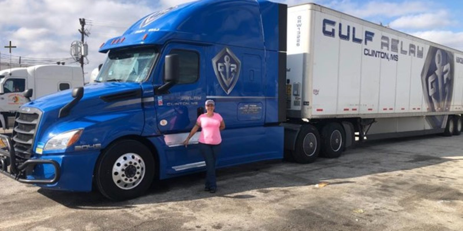 A person in a pink shirt and hat stands next to a large blue Gulf Relay semi-truck and trailer in a parking lot under a partly cloudy sky.