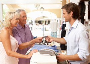 An older couple stands at a store counter, smiling as the man hands a credit card to a young male cashier. Clothing items and a card payment terminal are on the counter. The setting appears bright and welcoming.