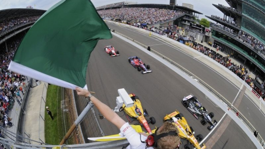 A race official waves a green flag to start an IndyCar race as several race cars speed past the starting line on a track, with a large crowd in the grandstands.