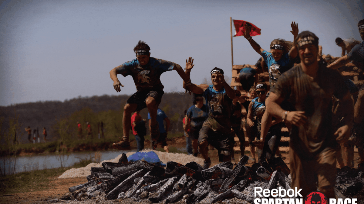A group of athletes wearing headbands and muddy clothes jump over flaming logs during a Reebok Spartan Race obstacle course outdoors. The event logo is visible in the bottom right corner.