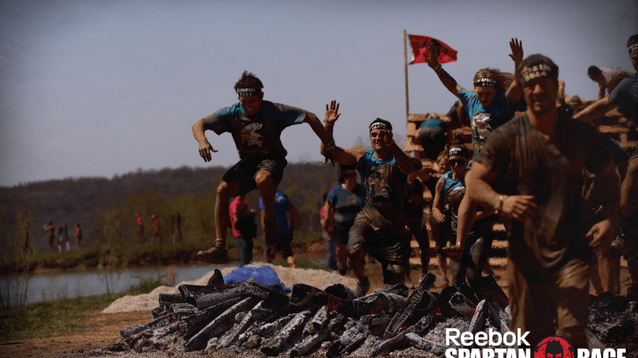 A group of athletes wearing headbands and muddy clothes jump over flaming logs during a Reebok Spartan Race obstacle course outdoors. The event logo is visible in the bottom right corner.