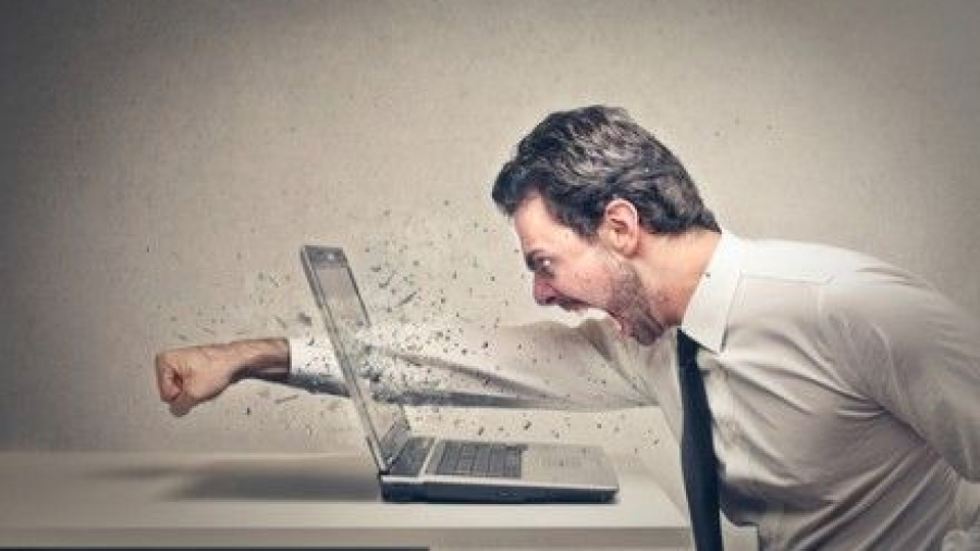 A man in a dress shirt and tie angrily punches a laptop on a desk, causing the screen to shatter and pieces to fly outward.