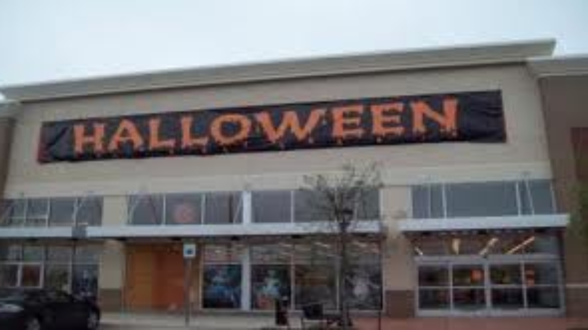 A large retail store exterior with a black banner reading HALLOWEEN in orange letters hanging above the entrance. The windows below display some decorations, and a small tree is in front of the building.