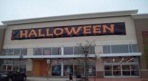 A large retail store exterior with a black banner reading HALLOWEEN in orange letters hanging above the entrance. The windows below display some decorations, and a small tree is in front of the building.