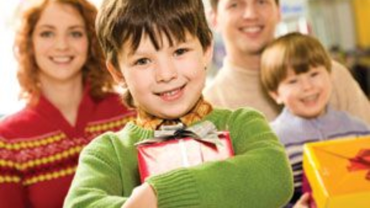 A smiling boy in a green sweater holds a wrapped gift, standing in front of a smiling family—an adult man, an adult woman, and another boy—each holding presents. The background is brightly lit indoors.