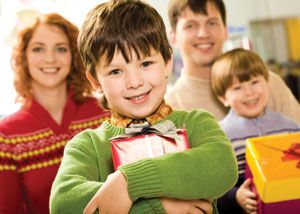 A smiling boy in a green sweater holds a wrapped gift, standing in front of a smiling family—an adult man, an adult woman, and another boy—each holding presents. The background is brightly lit indoors.