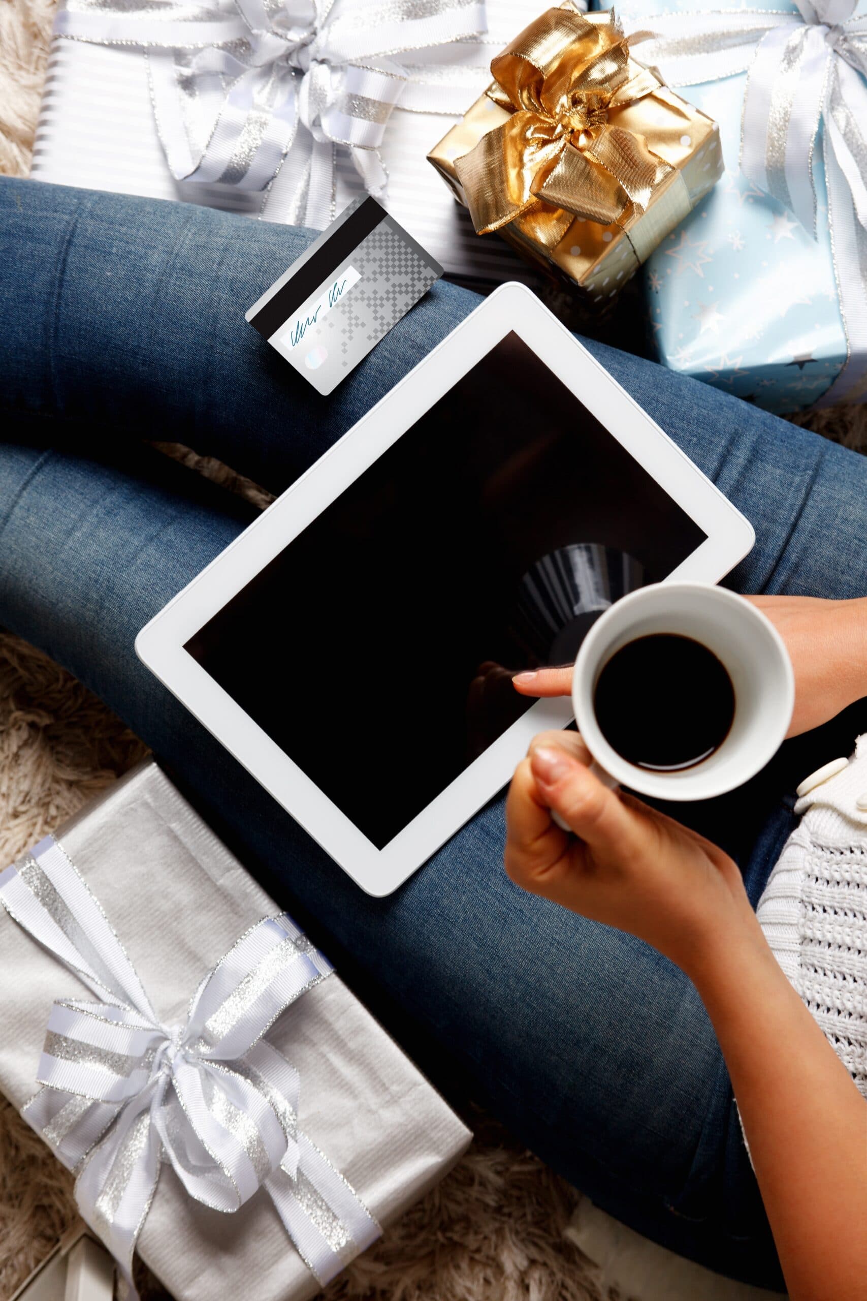 A person sitting with wrapped gifts holds a cup of coffee and a tablet, with a credit card nearby, suggesting online holiday shopping or gifting.