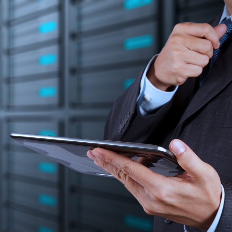 A man in a suit adjusts his tie while holding a tablet, standing in front of rows of server racks in a data center, highlighting the efficiency of goods-to-person technology in modern logistics operations.