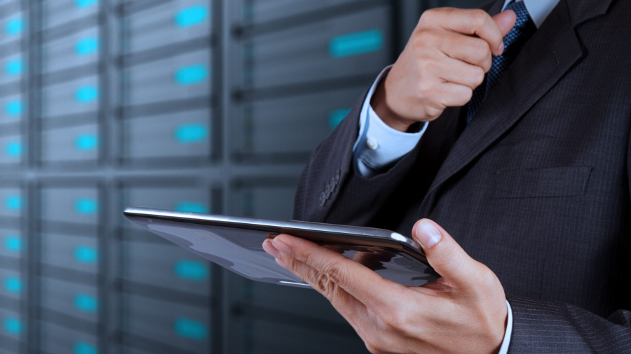 A man in a suit adjusts his tie while holding a tablet, standing in front of rows of server racks in a data center, highlighting the efficiency of goods-to-person technology in modern logistics operations.