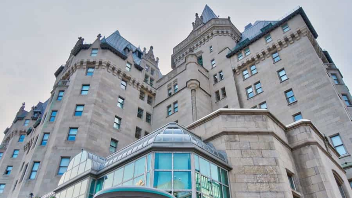 A view looking up at a large, historic stone building with turrets and steep blue-gray roofs, featuring many windows and a modern glass entrance in the foreground.