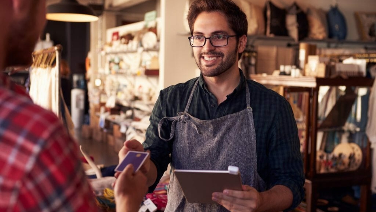 A smiling shop worker in an apron holds a tablet and processes a payment from a customer handing over a credit card in a cozy store environment. Shelves with various items are visible in the background.