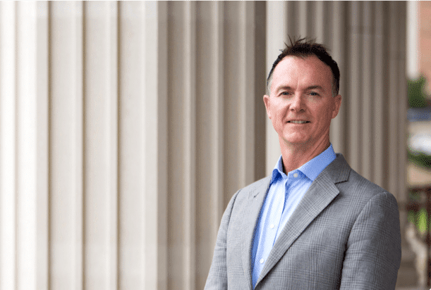 A man in a gray suit jacket and blue shirt stands in front of tall, white columns, smiling at the camera.