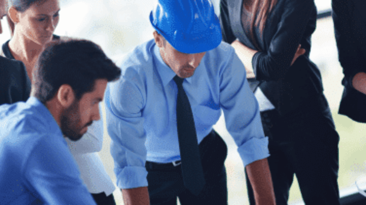 A group of business professionals and an engineer wearing a blue hard hat are gathered around a table, closely examining blueprints or architectural plans in a bright office setting.