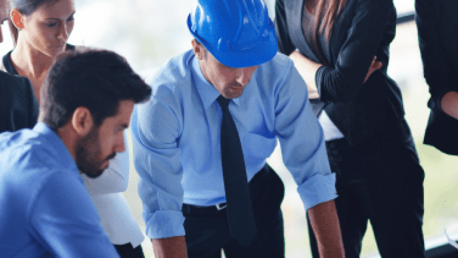 A group of business professionals and an engineer wearing a blue hard hat are gathered around a table, closely examining blueprints or architectural plans in a bright office setting.