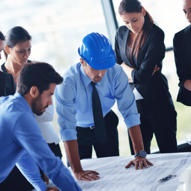 A group of business professionals and an engineer wearing a blue hard hat are gathered around a table, closely examining blueprints or architectural plans in a bright office setting.