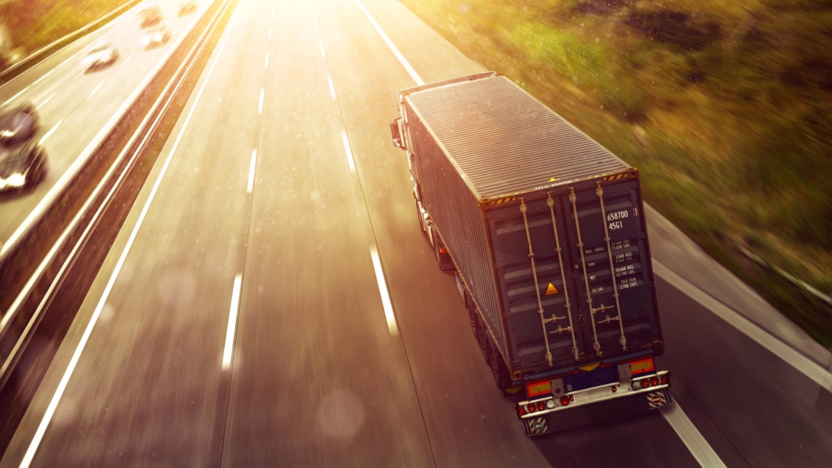 A container truck drives on a sunlit highway, surrounded by greenery, with a few cars visible in other lanes. The bright sunlight creates a warm, glowing effect over the road.