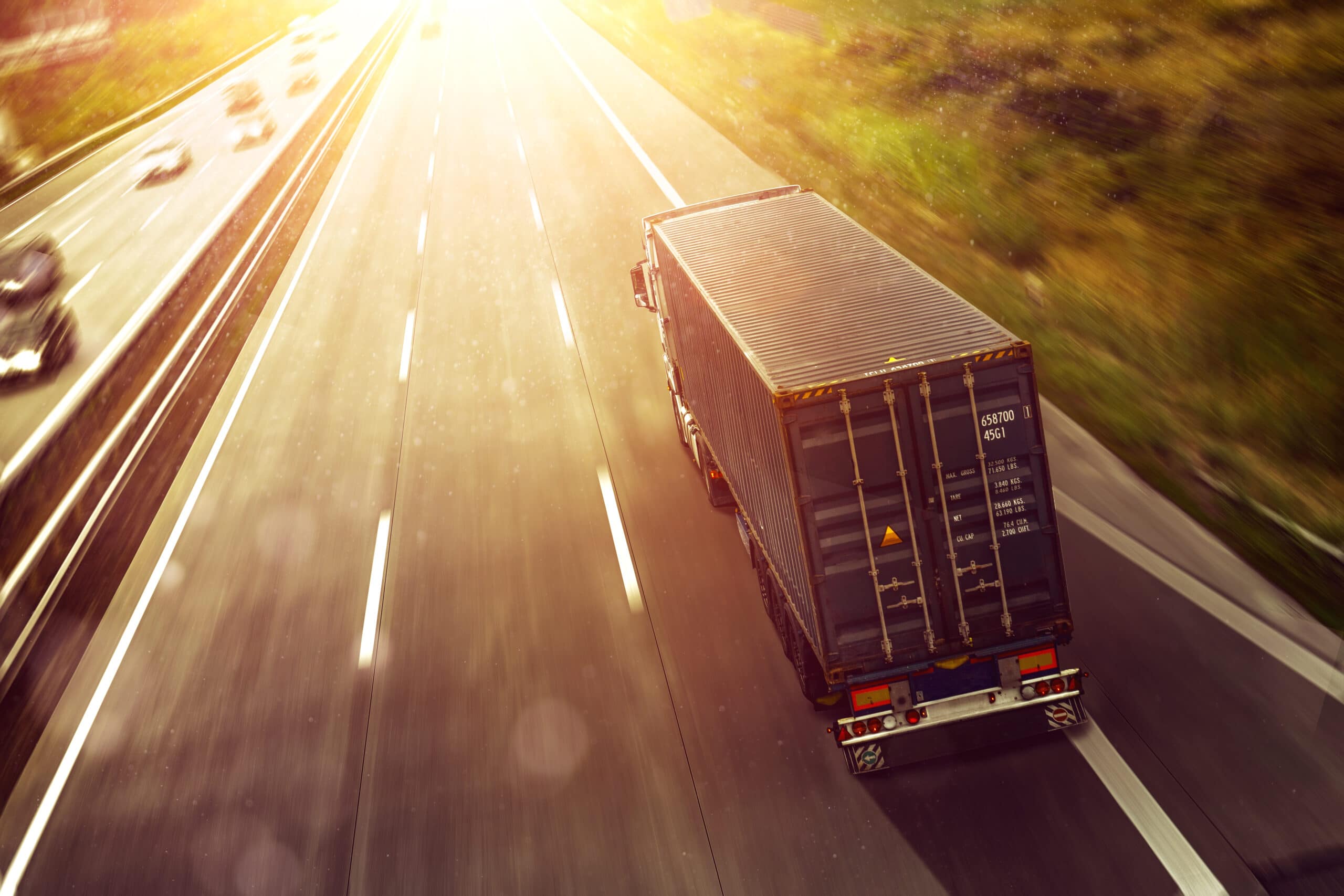 A container truck drives on a sunlit highway, surrounded by greenery, with a few cars visible in other lanes. The bright sunlight creates a warm, glowing effect over the road.