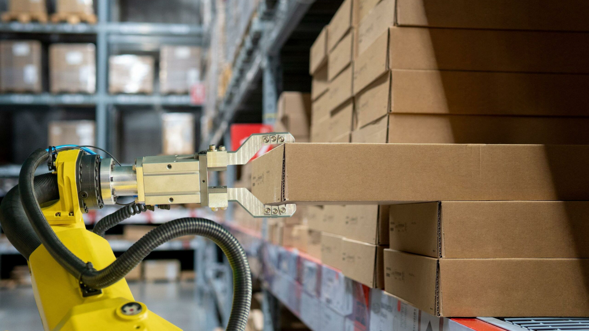 A robotic arm with a metal gripper lifts a cardboard box from a shelf in a dark warehouse filled with stacked boxes and shelves.