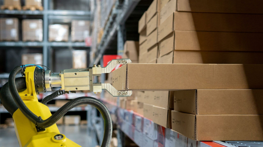 A robotic arm with a metal gripper lifts a cardboard box from a shelf in a dark warehouse filled with stacked boxes and shelves.