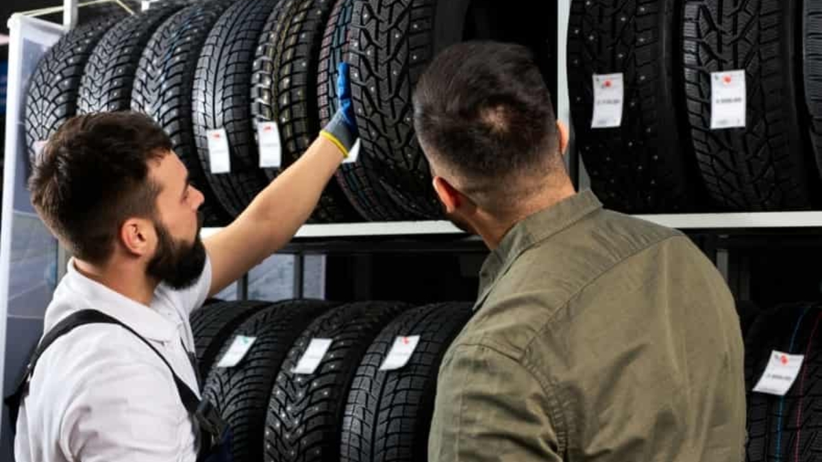 Two men examine and discuss tires displayed on shelves in a store; one man wearing a glove points at a tire while the other listens, with rows of tires and price tags visible.