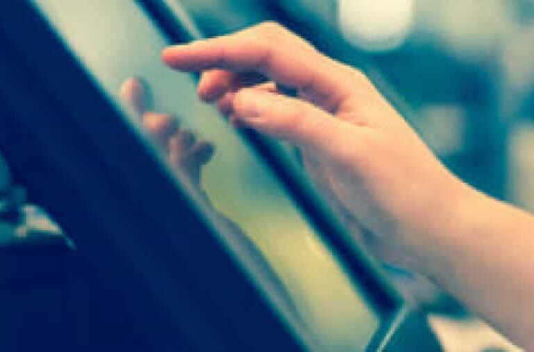 A close-up of a hand touching the screen of a touchscreen device, likely a kiosk or tablet, with a blurred background.