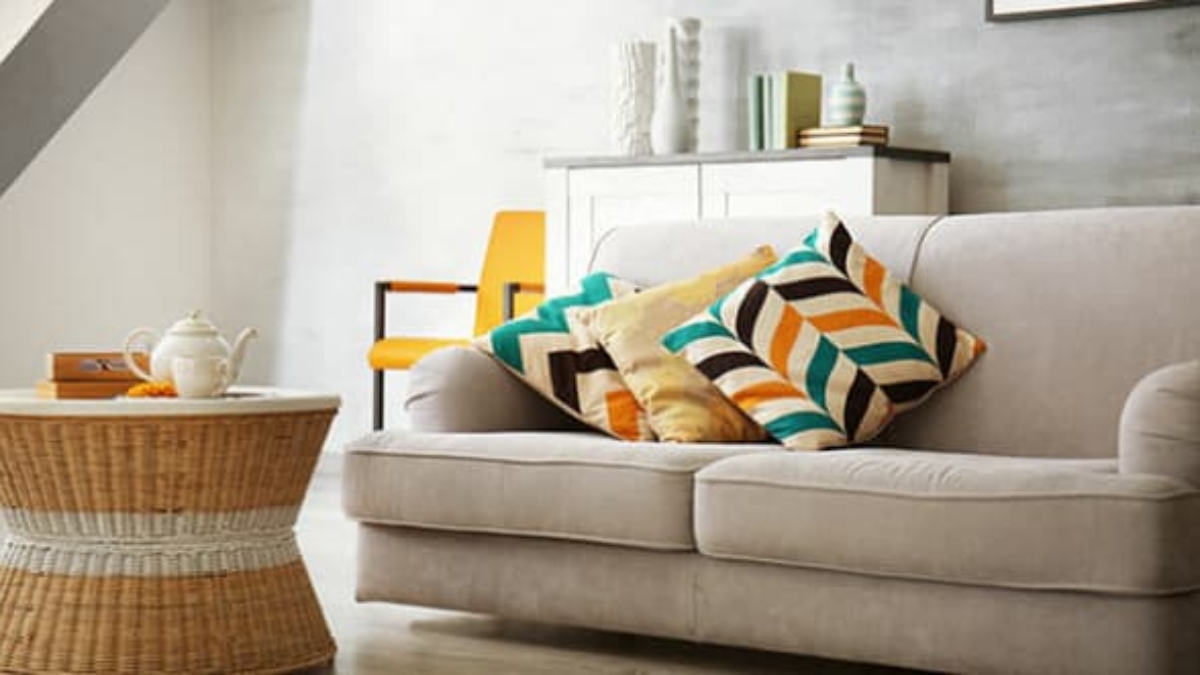 A beige sofa with colorful patterned cushions beside a round wicker coffee table with a teapot set. In the background, a white cabinet and an orange chair add accents to the modern, bright living room.
