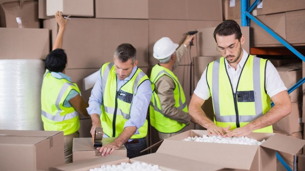 Four workers in high-visibility vests pack and organize cardboard boxes in a warehouse. One fills a box with packing material, another tapes a box, while others arrange boxes on shelves.