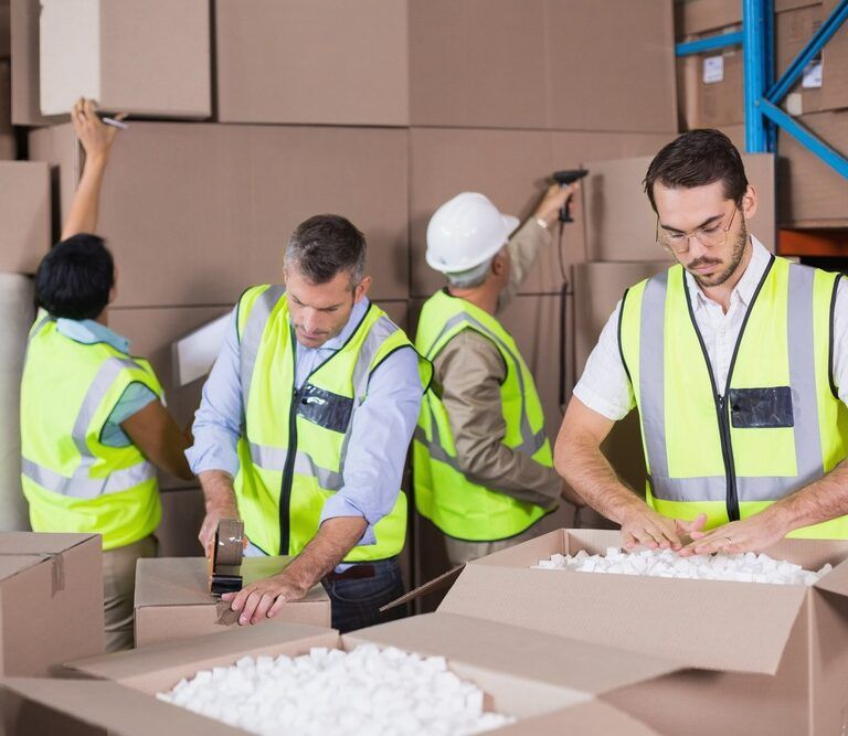 Four workers in high-visibility vests pack and organize cardboard boxes in a warehouse. One fills a box with packing material, another tapes a box, while others arrange boxes on shelves.