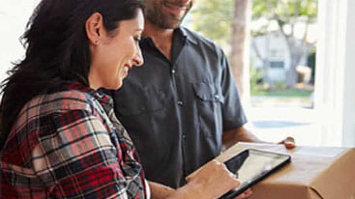 A woman smiles as she signs for a package on a digital tablet held by a delivery person standing beside her.