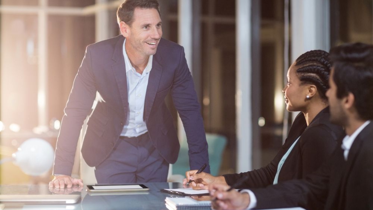 A man in a blue suit stands and smiles while speaking to two colleagues seated at a conference table with notebooks and a tablet in a modern office setting.